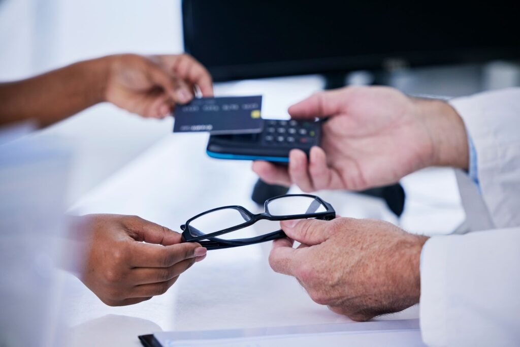 Close-up of two pairs of hands. One holds a pair of glasses and a payment terminal, while the other holds a credit card.