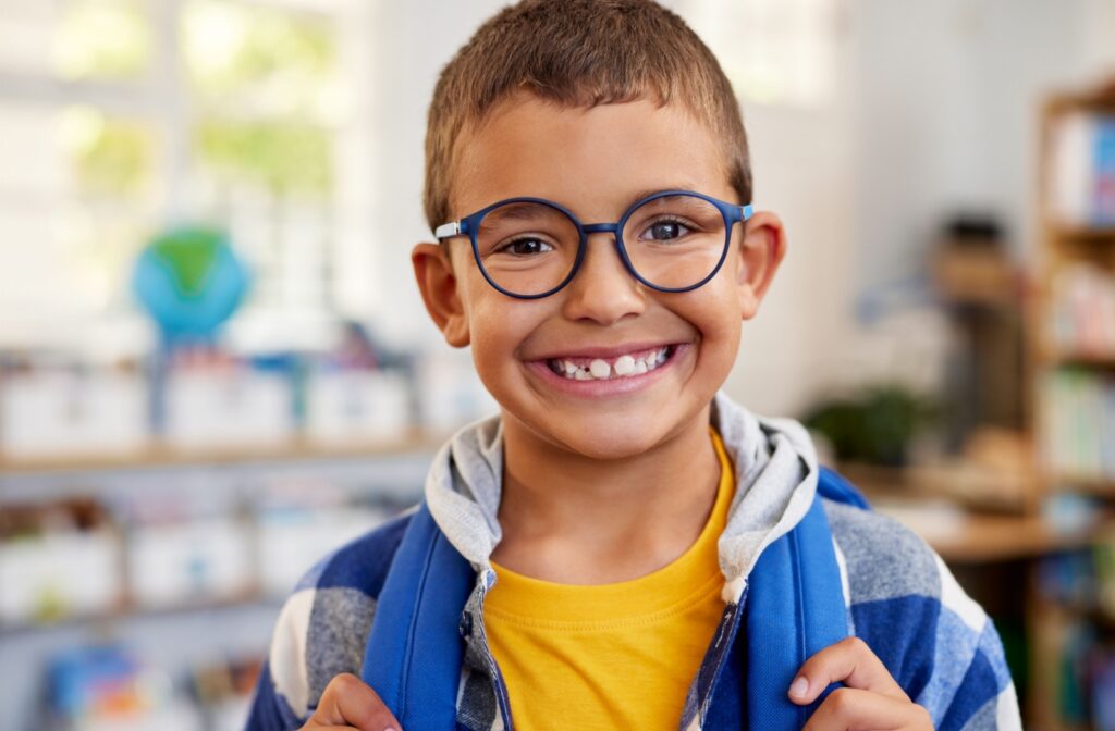 A child with glasses smiles at the camera, clutching his backpack straps.