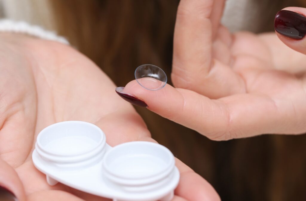 Close-up of a white contact lens case in one hand and a contact lens on a finger in another hand.