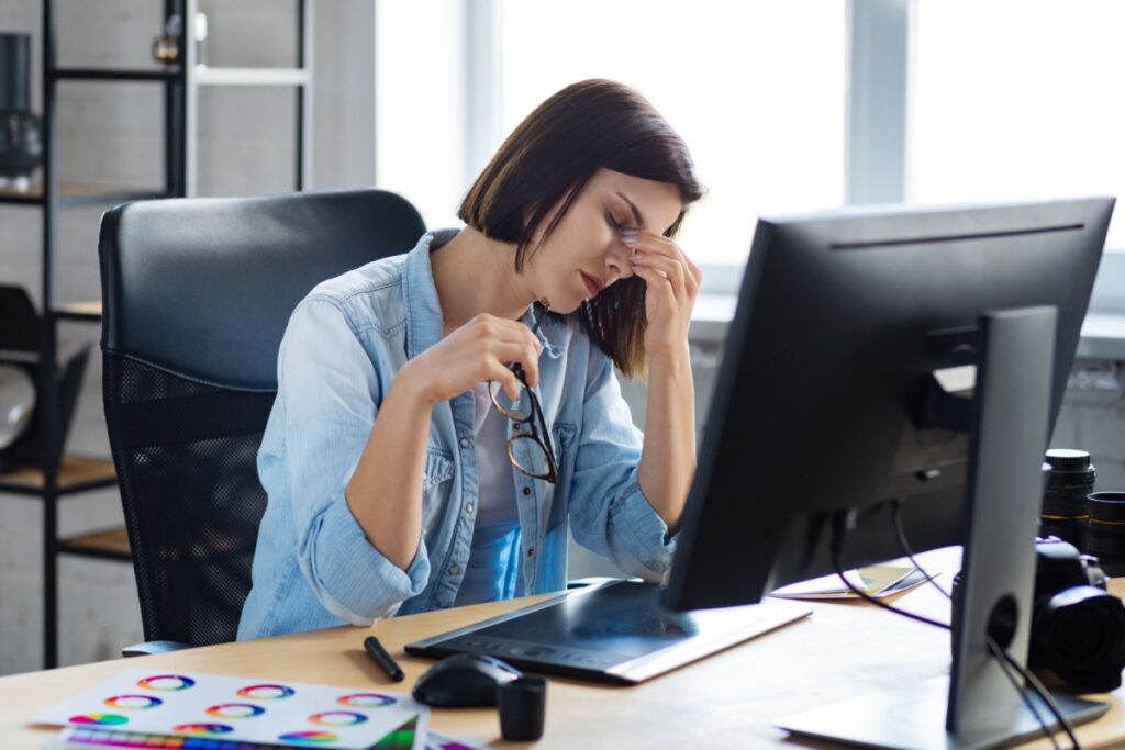 A woman sitting at her desk in front of a computer, rubbing her eyes and holding her glasses, showing signs of digital eye strain from prolonged screen use.