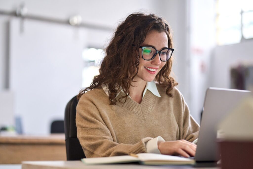 A woman wearing glasses and a beige sweater smiling while working on her laptop, representing comfortable vision and reduced eye strain during computer use.