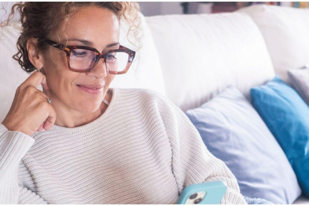 A woman with curly hair wearing stylish, tortoise-shell reading glasses, comfortably seated on a sofa while smiling and looking down at her smartphone.